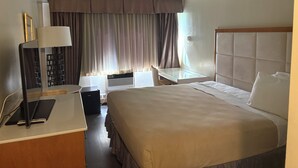 A hotel room with a bed, bedside table, lamp, and a view of the outdoors through curtains.