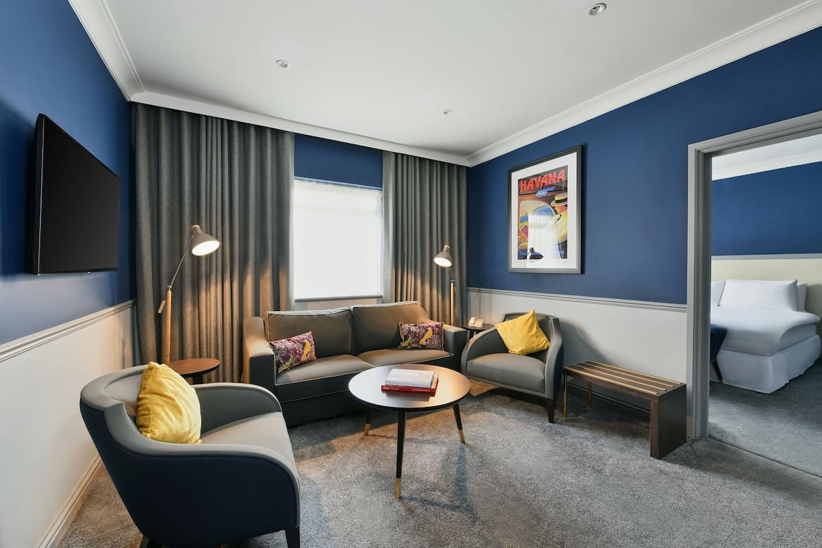Una habitación de hotel moderna con una pared azul oscuro de acento, un televisor de pantalla plana, un conjunto de sofás, una mesa de centro y un banco.