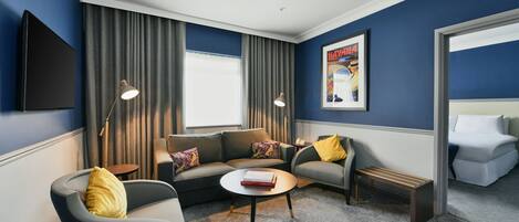 A modern hotel room with a dark blue accent wall, a flat-screen TV, a sofa set, a coffee table, and a bench.