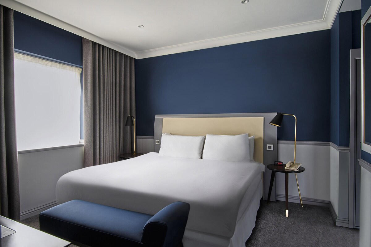 A hotel room with a large bed, a bedside table with a lamp, a small desk, and a window with curtains.