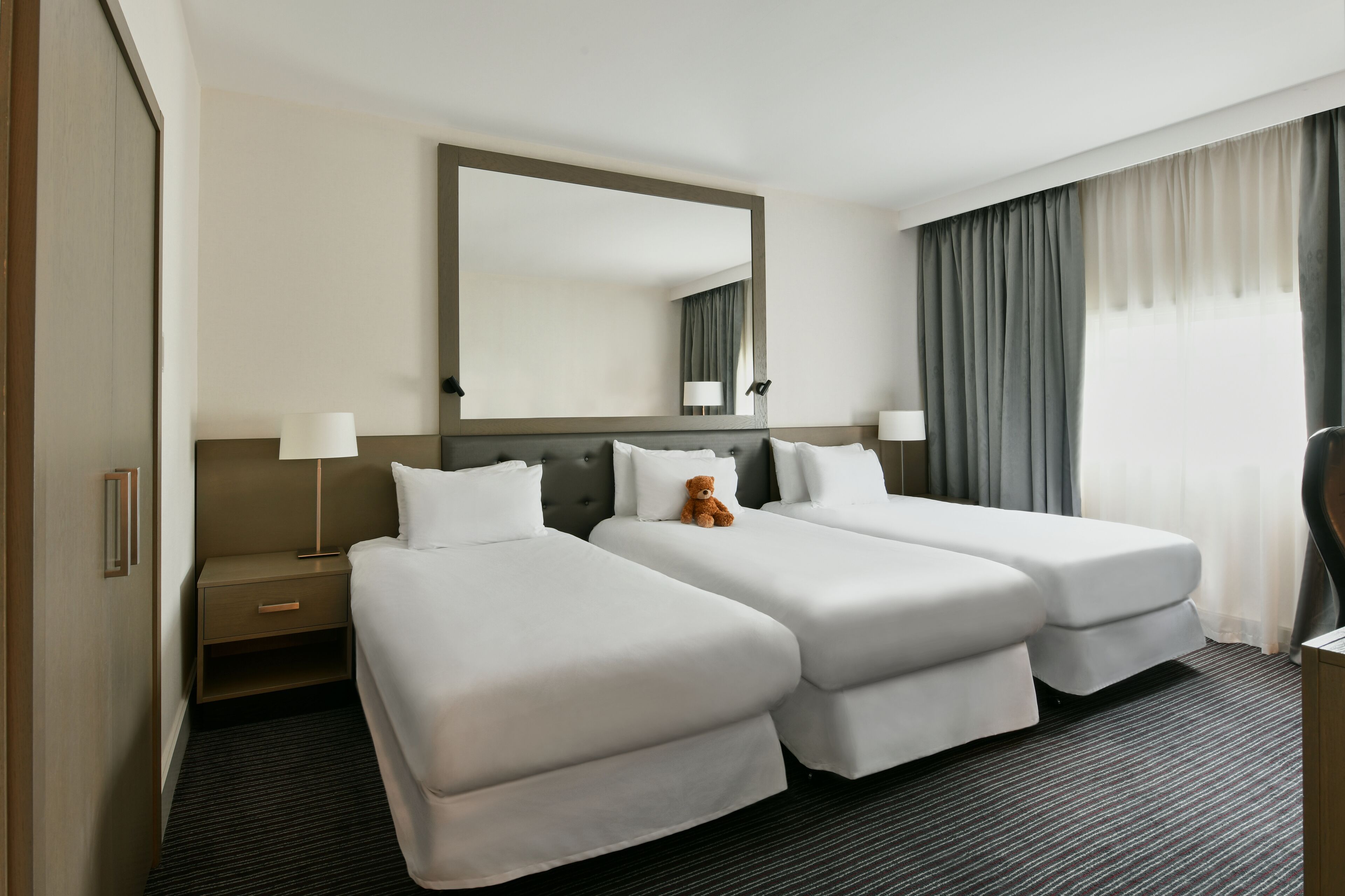 Habitación de hotel con dos camas individuales, un espejo grande, una mesita de noche con lámpara y vistas al exterior a través de una ventana con cortinas.