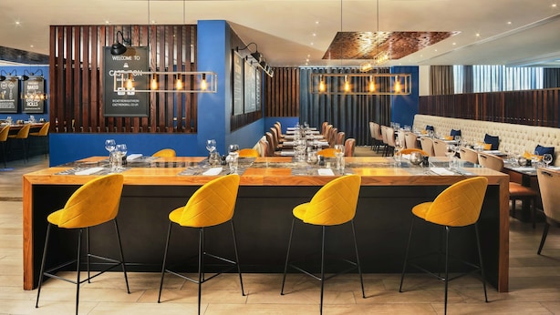A modern restaurant with a central bar area, wooden ceiling, and yellow bar stools.