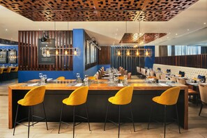 Ein modernes Restaurant mit einer zentralen Bar, Holzbalkendecke und gelben Barhocker.