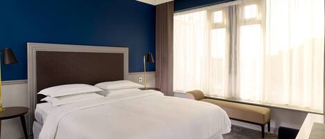 A hotel room with a large bed, a bedside table, a lamp, a bench, and a window with curtains.