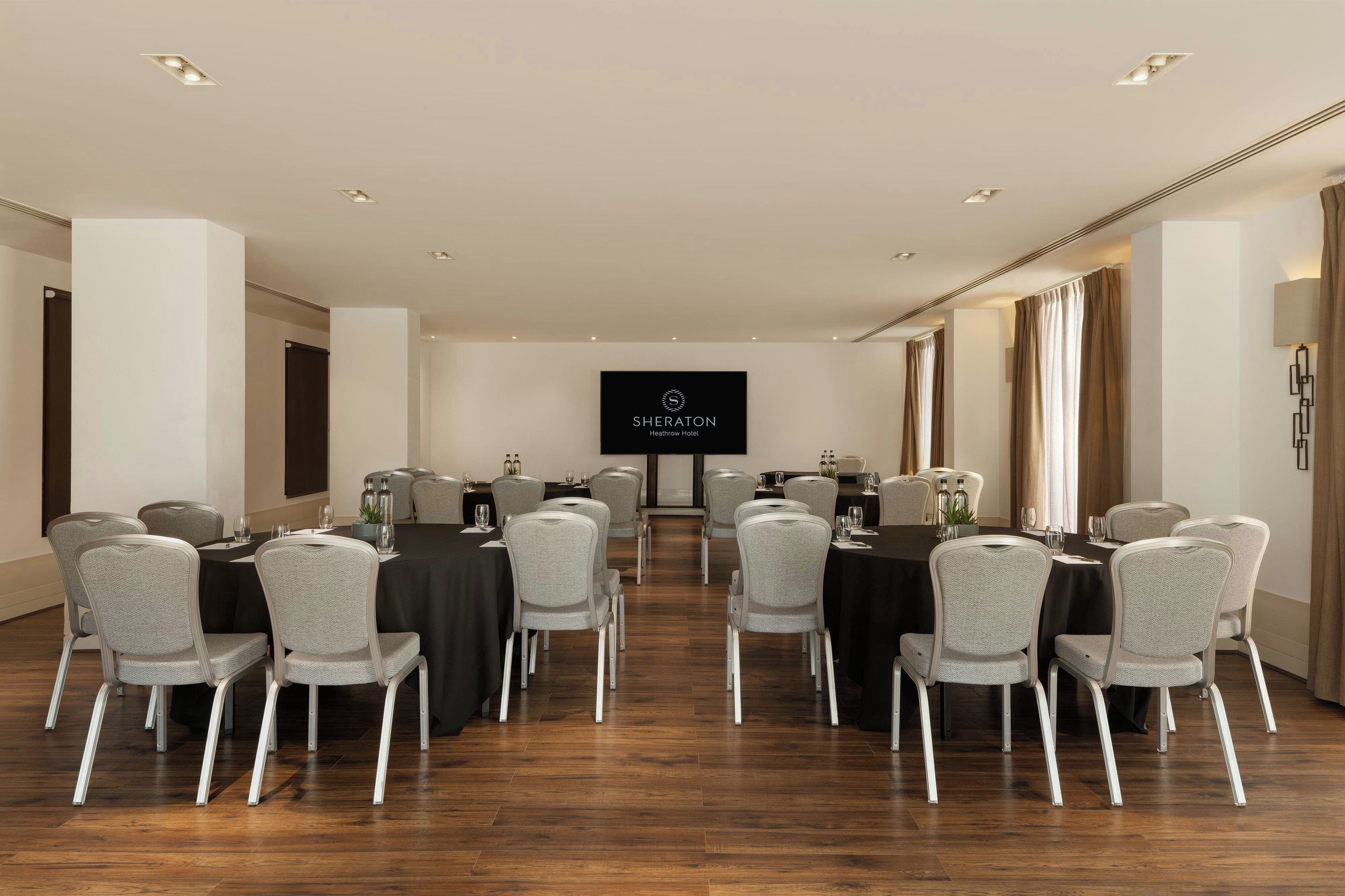 A conference room with a large screen displaying 'SHERATON Heathrow Hotel' logo, arranged tables with chairs, and a wooden floor.
