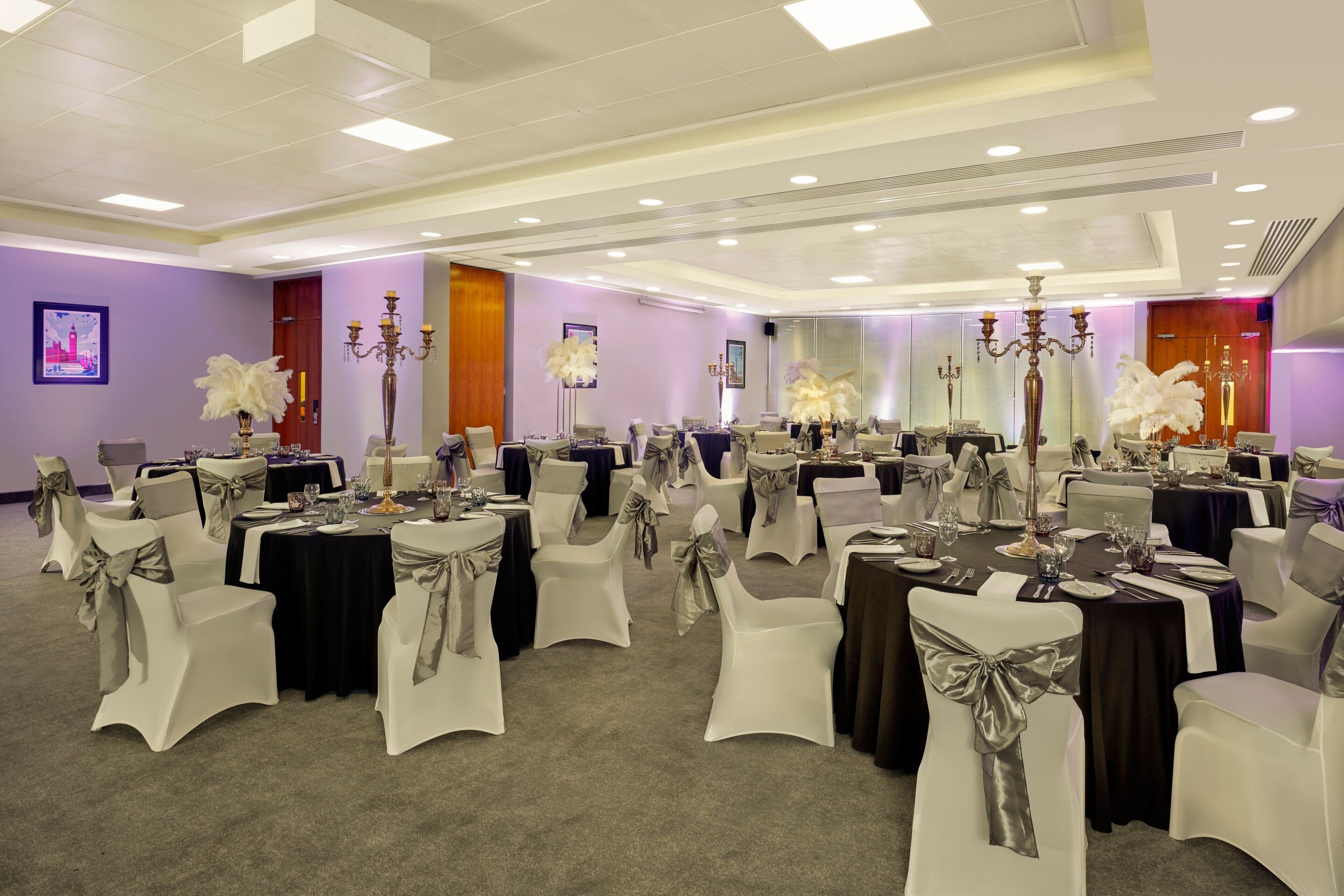 Een feestzaal met ronde tafels, witte stoelen met zilveren linten en kandelaars.