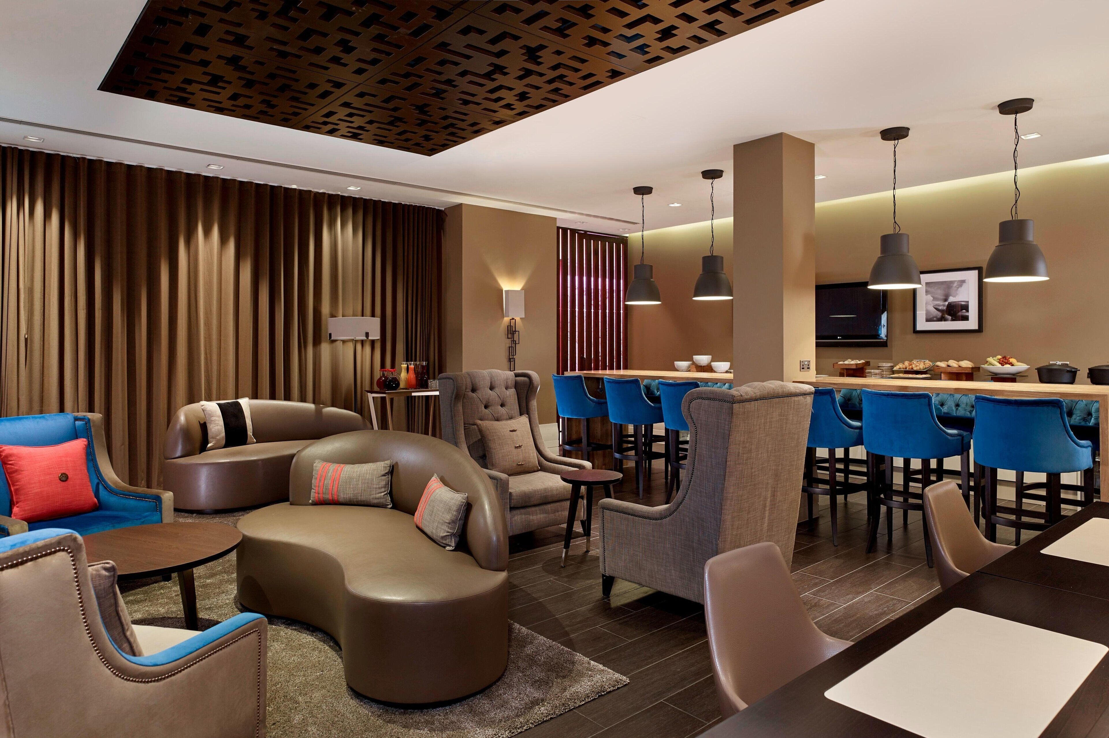 A modern hotel lobby with a curved sofa, armchairs, a dining area with blue chairs, and pendant lighting.
