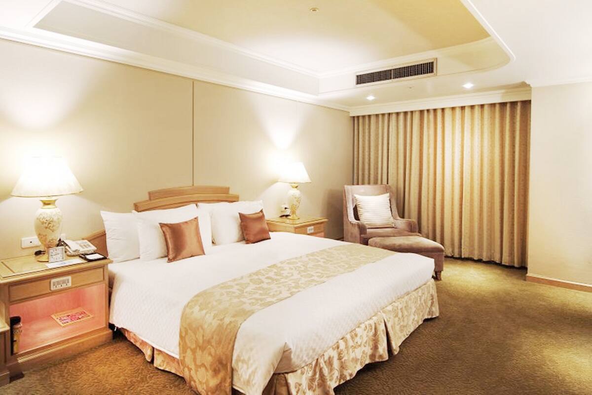 A hotel room with a large bed, two bedside tables, a chair, a window with curtains, and a ceiling vent.