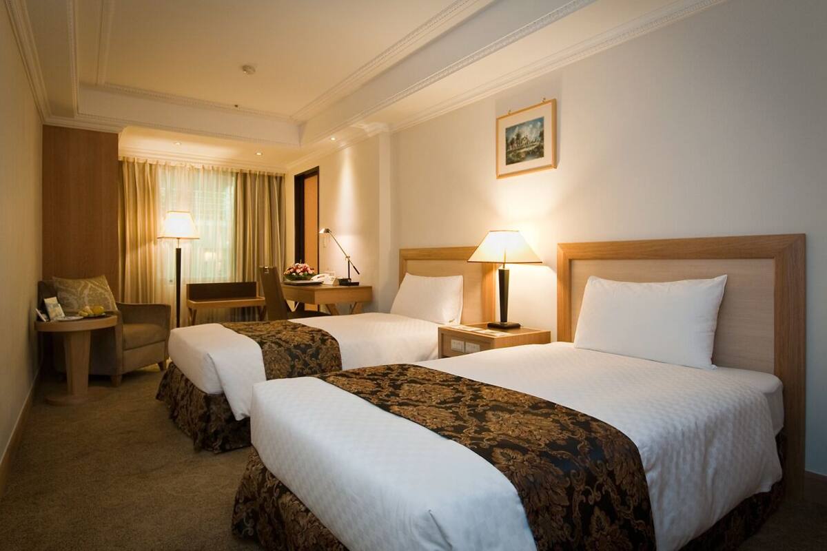 A hotel room with two beds, a desk, a chair, and a lamp.