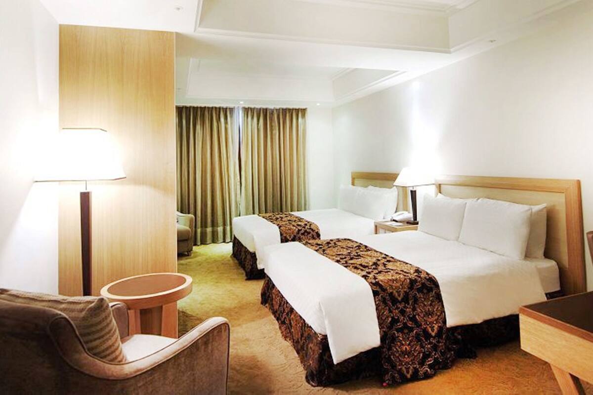 A hotel room with two beds, a desk, a chair, and a lamp.