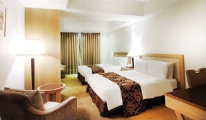 A hotel room with two beds, a desk, a chair, and a lamp.