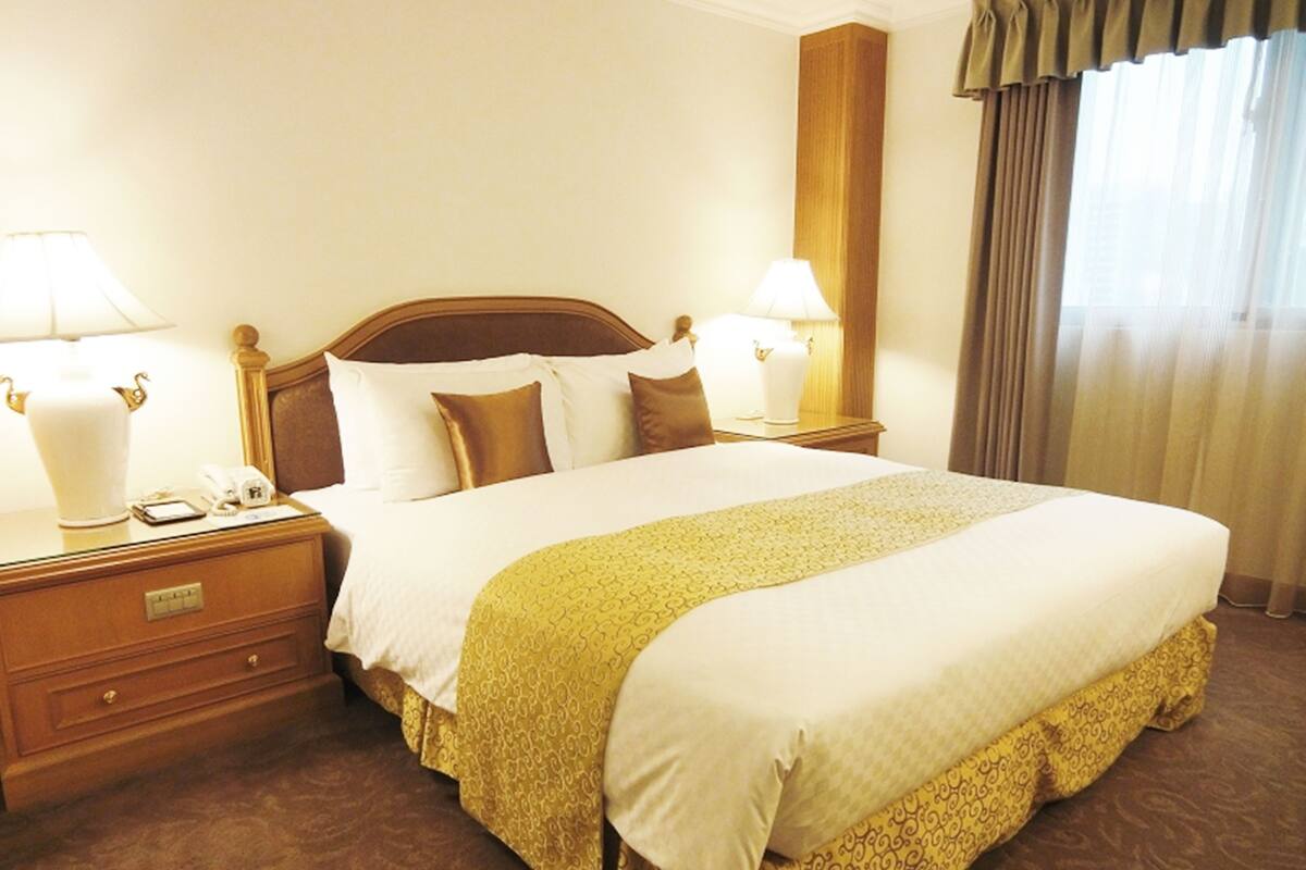 A hotel room with a large bed, two bedside tables with lamps, a wooden headboard, and a window with curtains.