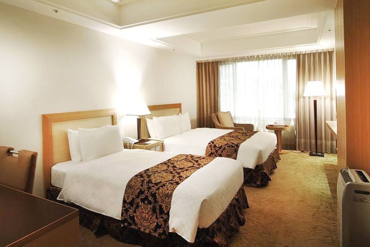 A hotel room with two beds, a desk, a chair, a lamp, and a window with curtains.