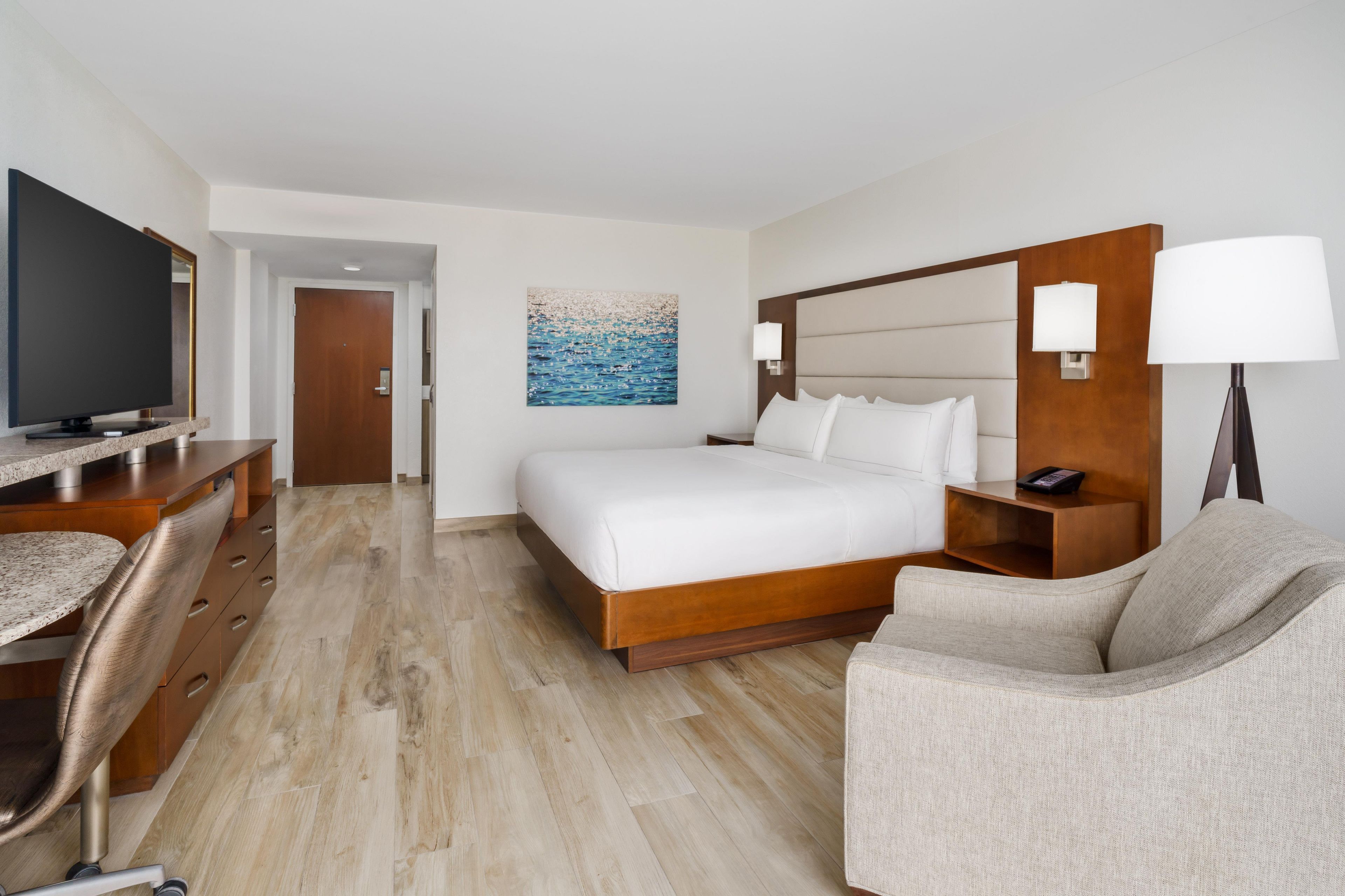A hotel room with a bed, a desk, a chair, a TV, and a painting on the wall.