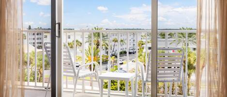 Un balcon avec des chaises blanches, une table et une vue sur un quartier résidentiel, avec des palmiers et des voitures garées.