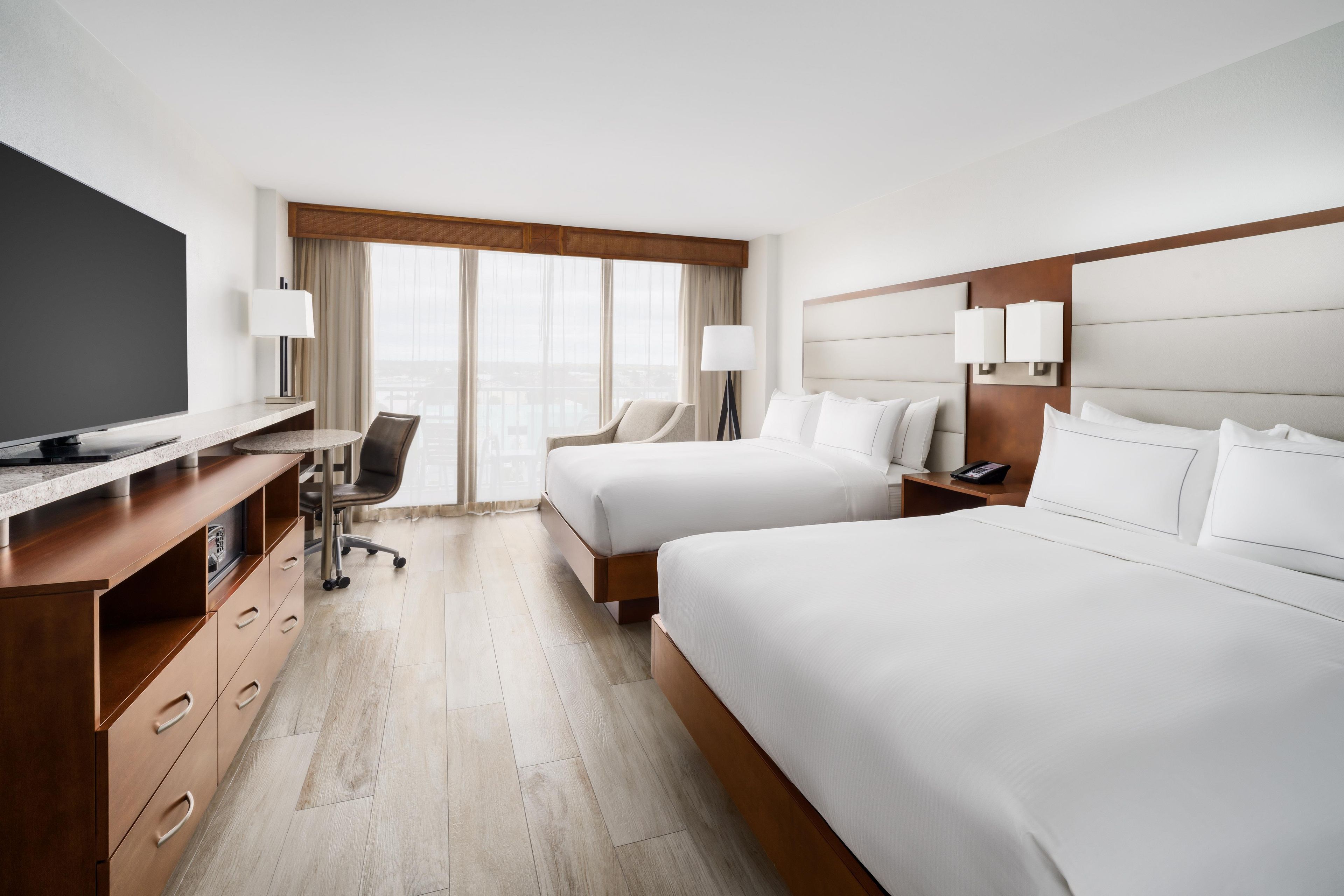 A hotel room with a TV, desk, two beds, a chair, and a view of the city.