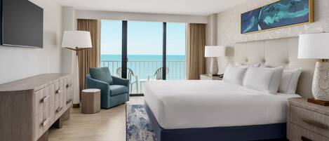 A hotel room with a large bed, a desk, a chair, a small side table, and a balcony with a view of the sea.