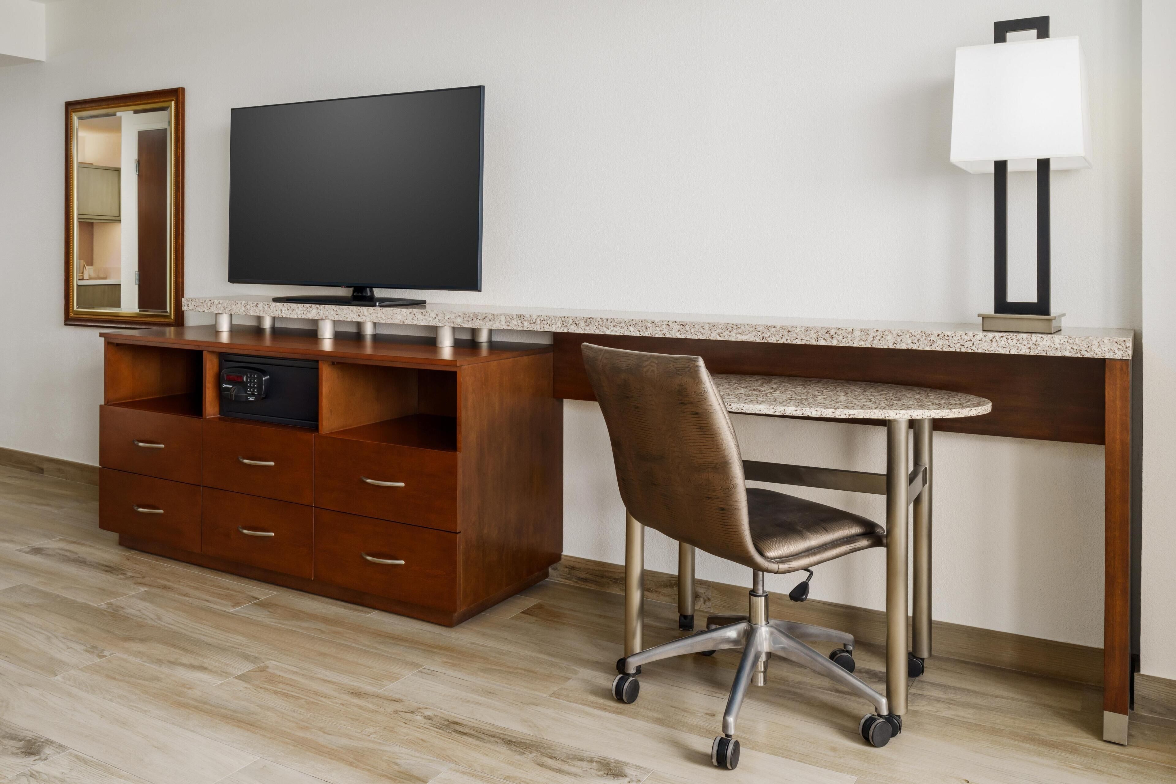 Una habitación de hotel moderna con un escritorio de madera, un televisor de pantalla plana, una silla, una lámpara y un mueble empotrado para televisión.