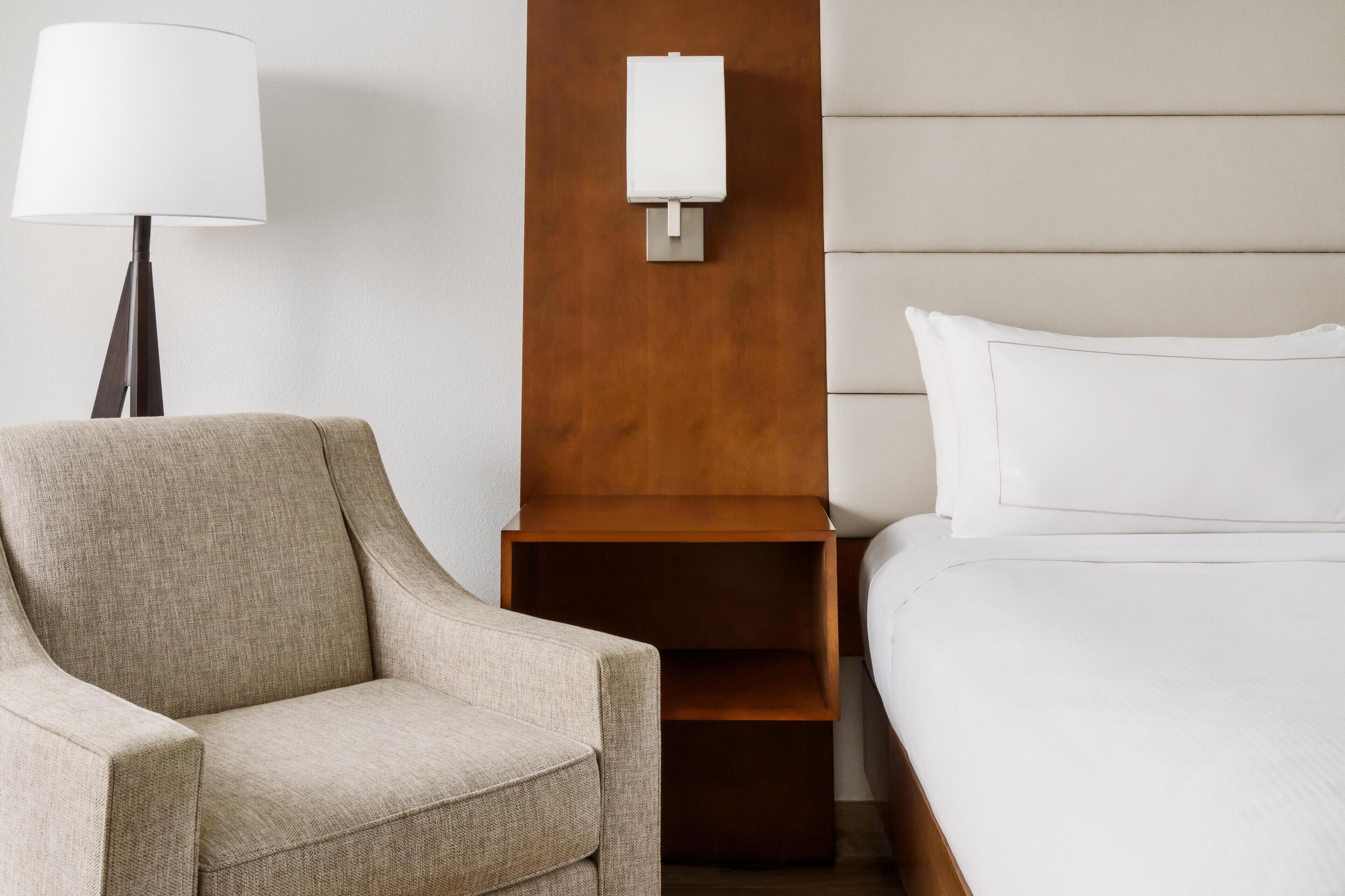 A hotel room with a beige armchair, a bedside table with a lamp, a wooden panel, and a bed with white bedding.