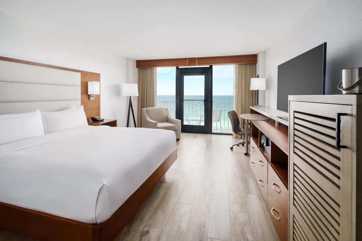 A hotel room with a large bed, a desk, a chair, a TV, and a view of the ocean.
