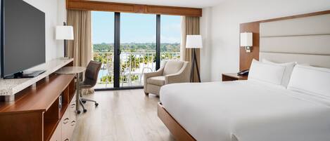 A hotel room with a large bed, a desk with a flat-screen TV, a chair, and a balcony with a view of the landscape.