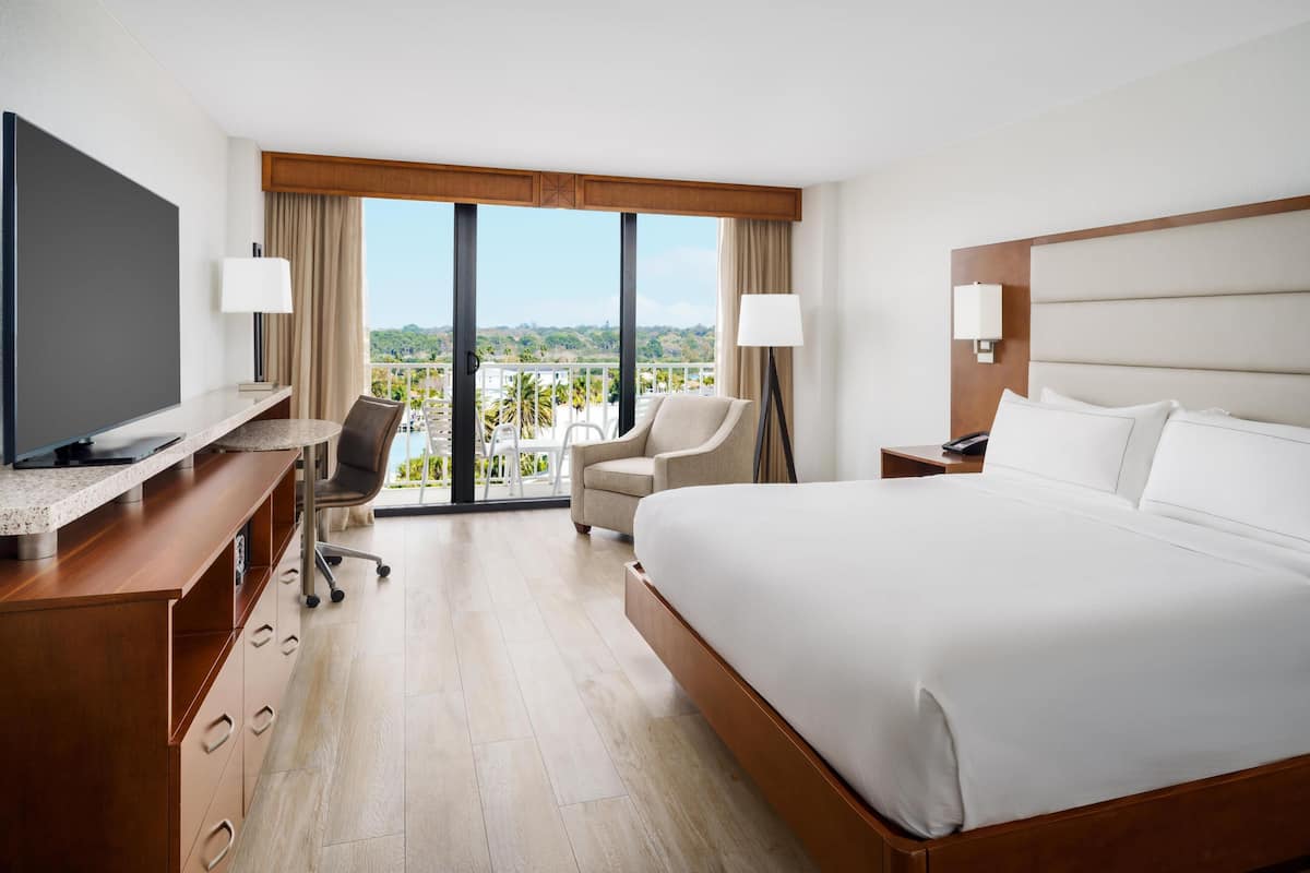 A hotel room with a large bed, a desk with a TV, a chair, and a balcony with a view.