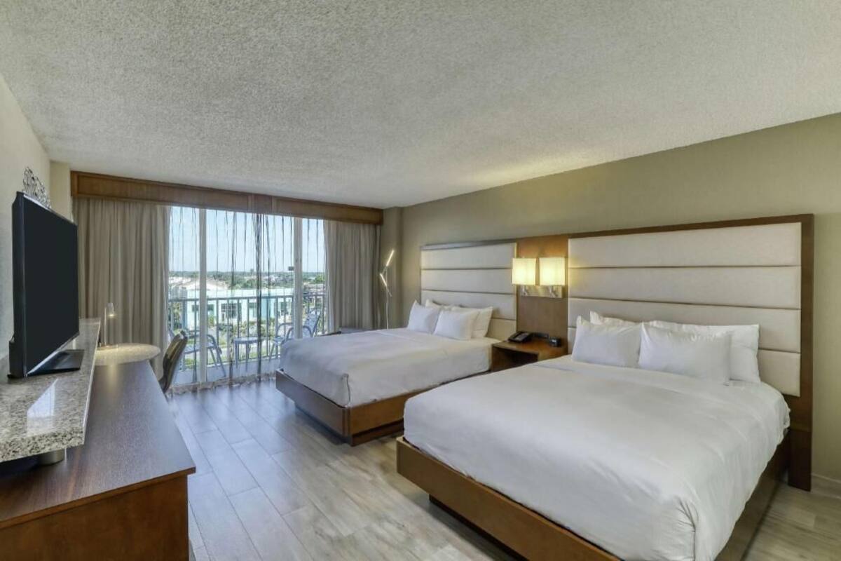 A hotel room with two beds, a desk, a TV, and a balcony with a view.