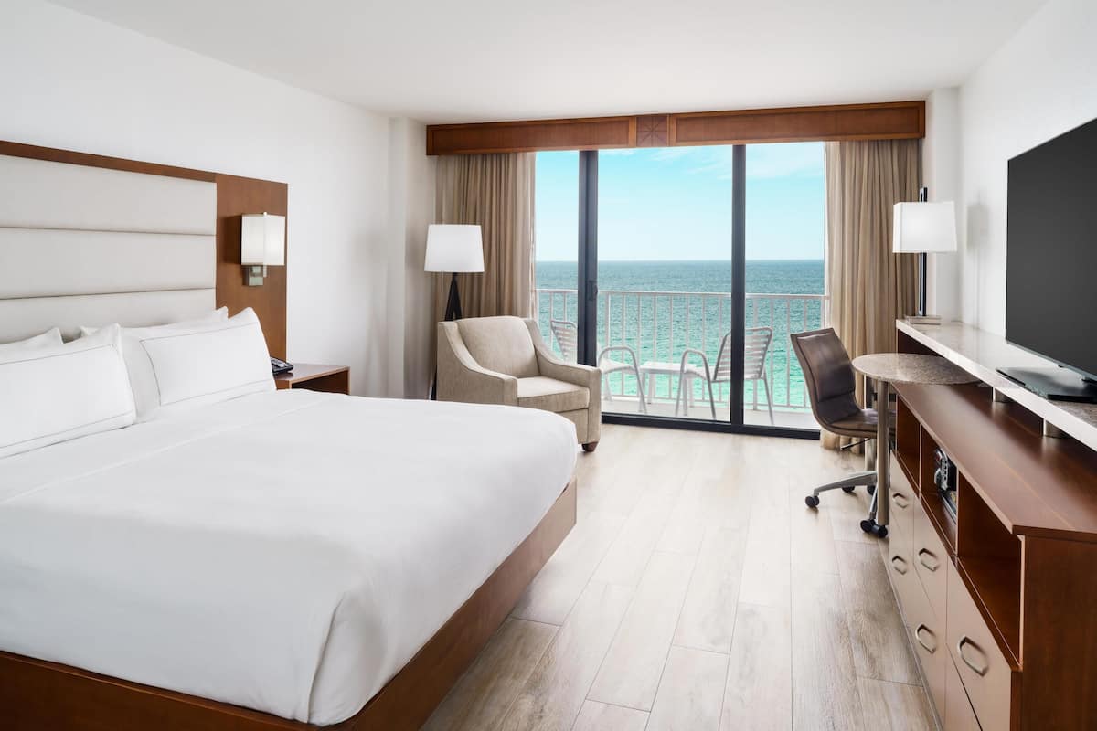 A hotel room with a large bed, a desk, a chair, a TV, and a balcony with a view of the ocean.