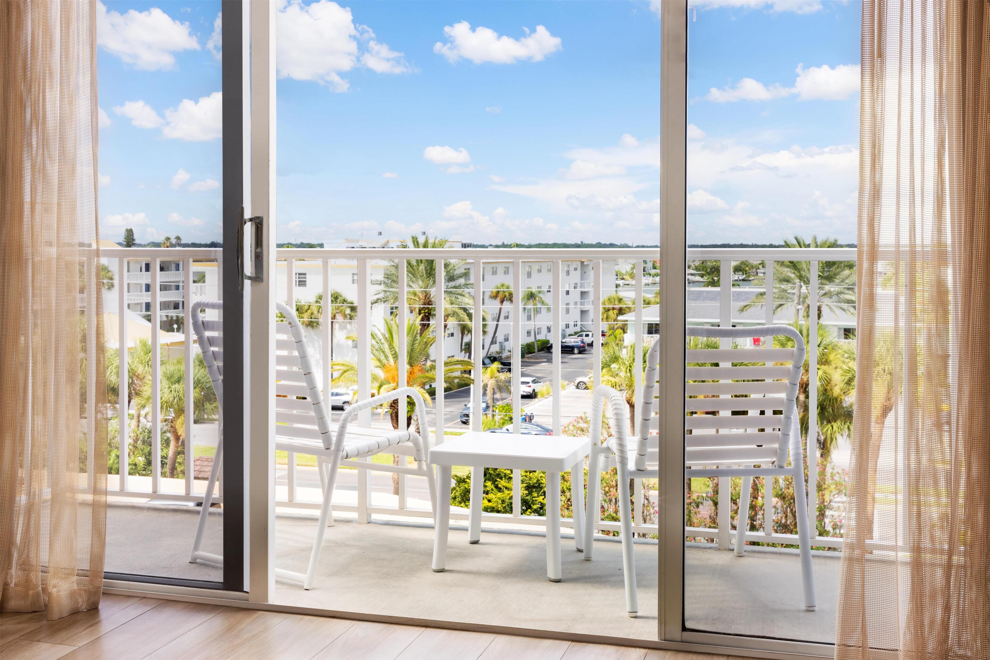 Ein Balkon mit weißen Stühlen und einem Tisch, der auf ein Wohngebiet mit Palmen und Gebäuden blickt.