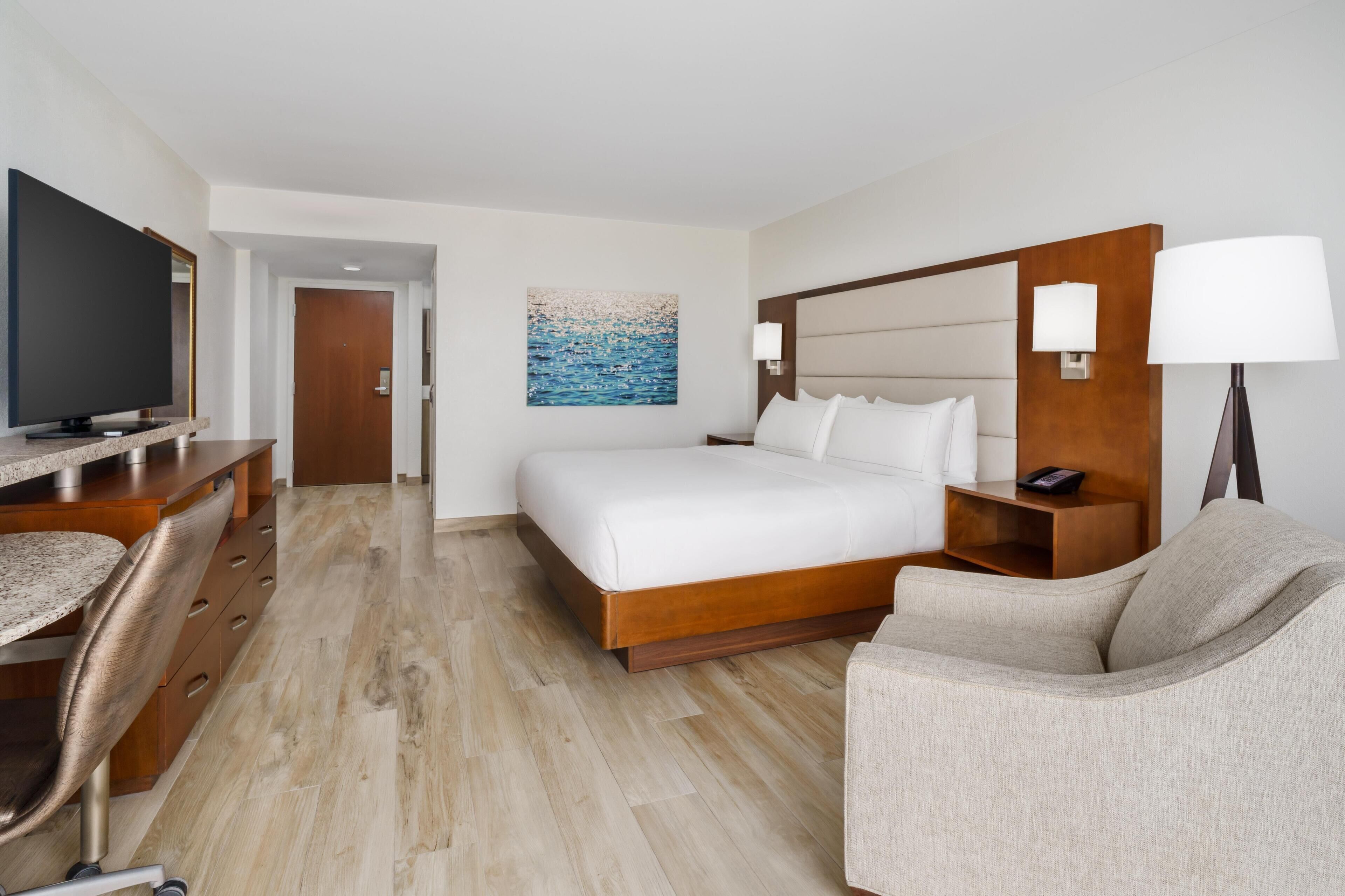 A hotel room with a bed, a desk with a television, a chair, a painting on the wall, and a lamp.