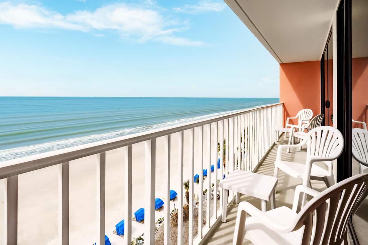 A balcony with white chairs and tables, overlooking a beach with a clear view of the ocean.