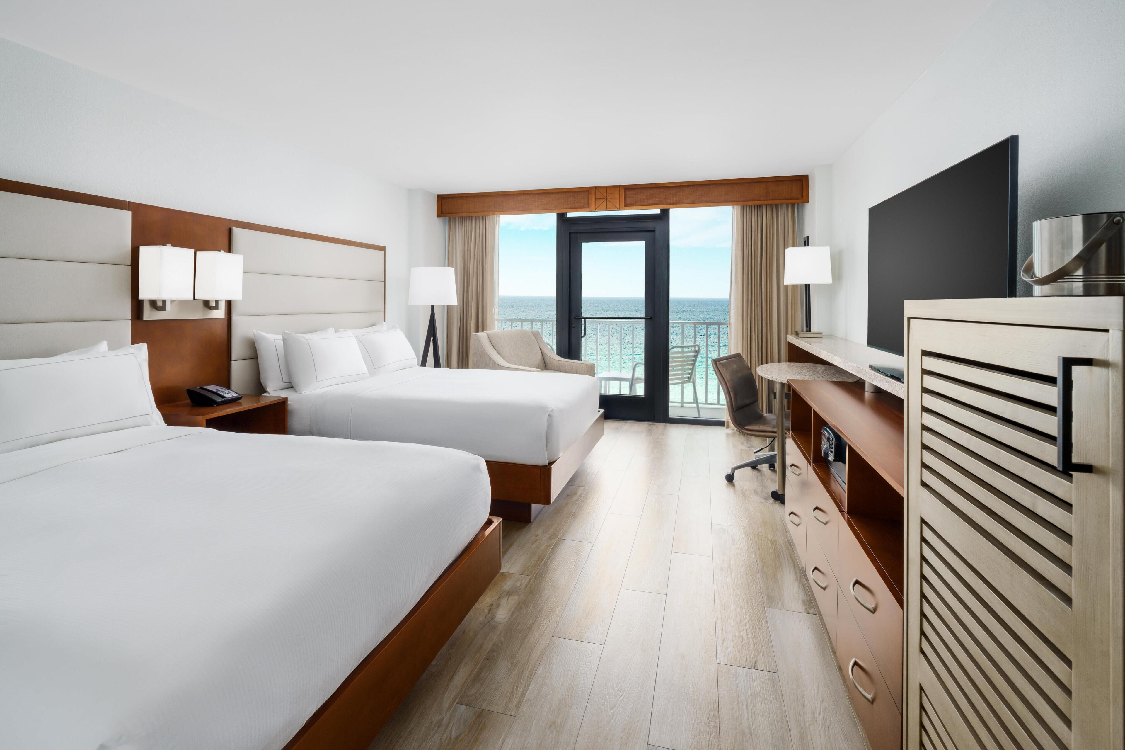 A hotel room with two beds, a desk, a TV, and a balcony with a view of the sea.