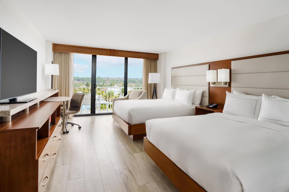 A hotel room with two beds, a desk, a TV, and a view of the city.