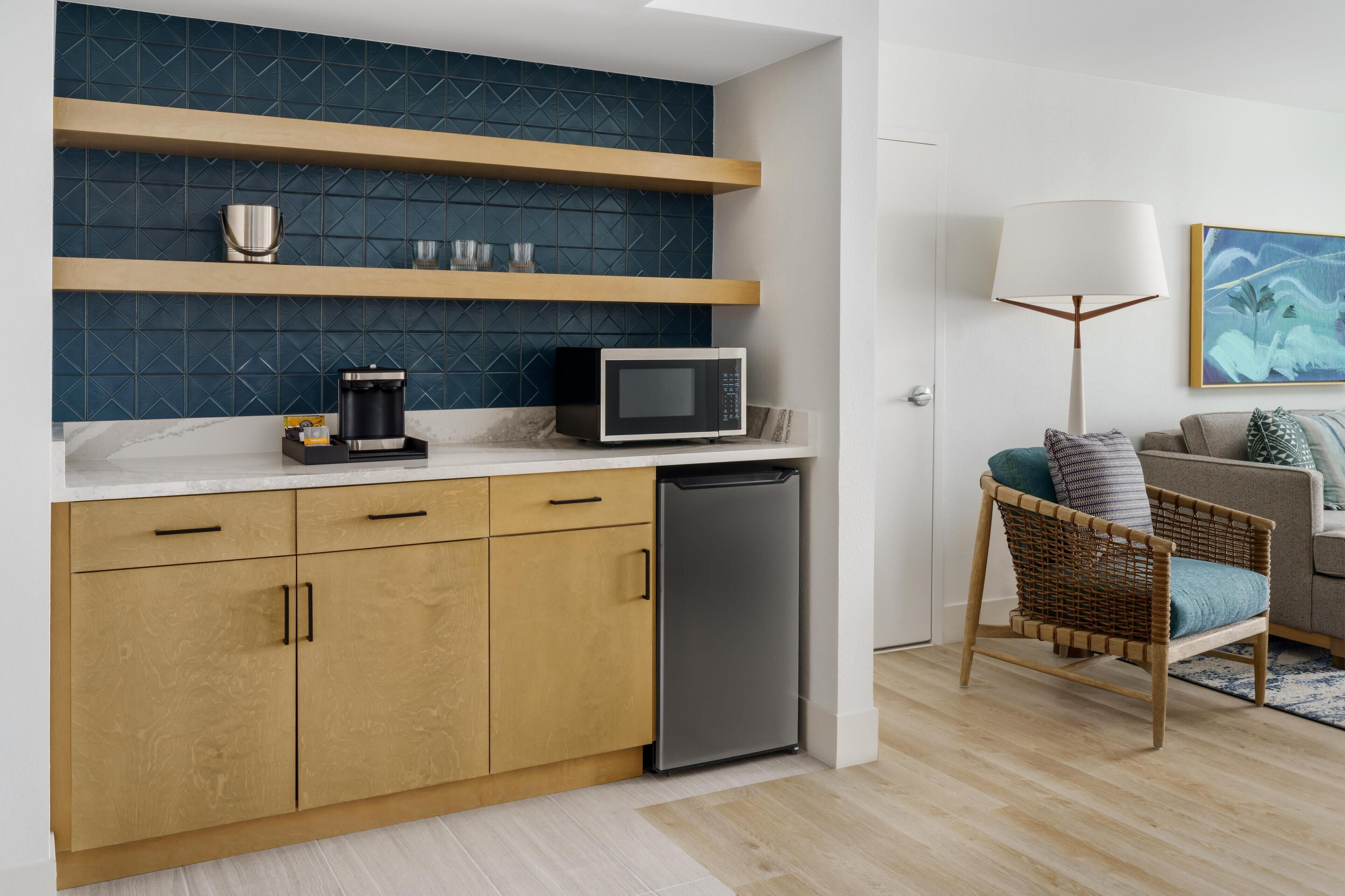 A compact kitchen with a microwave, coffee maker, and dishwasher. There is a wicker chair and a lamp in the adjacent living area.