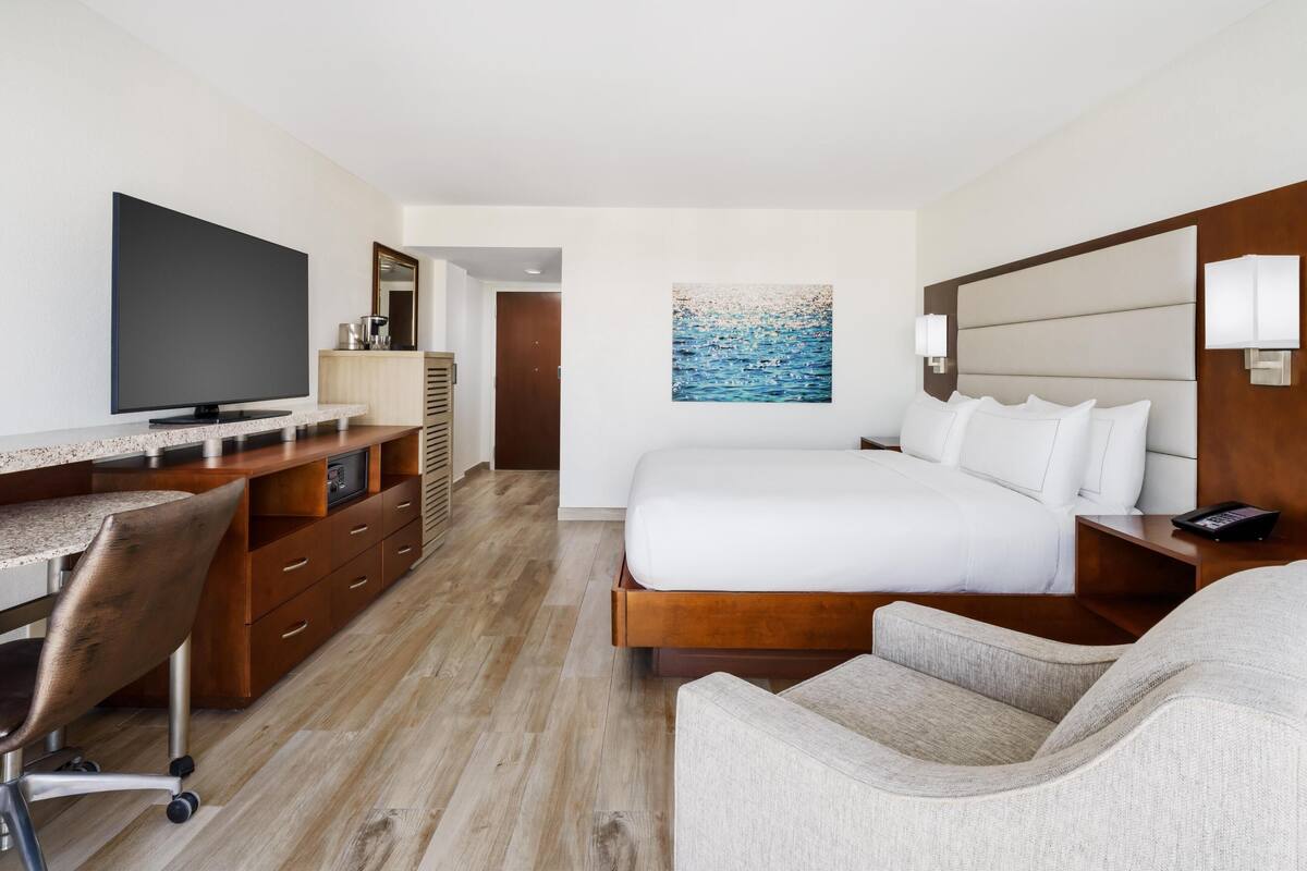 A hotel room with a bed, a desk with a flat-screen TV, a chair, a painting on the wall, and a wooden headboard.