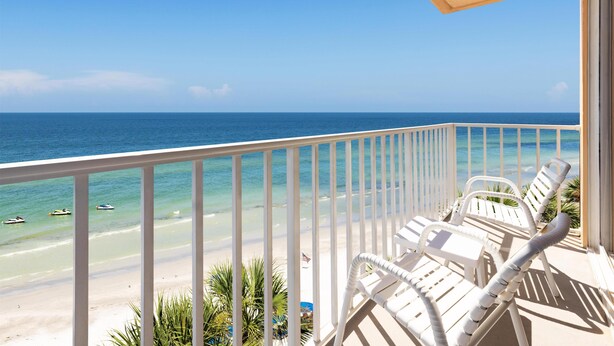Ein Balkon mit weißen Stühlen mit Blick auf einen Strand und klares, blaues Wasser sowie einige Boote.