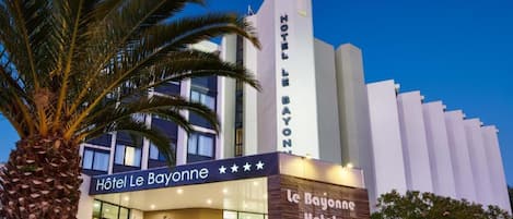The exterior of the Hôtel Le Bayonne, featuring a modern facade with a prominent vertical sign and a palm tree in the foreground.