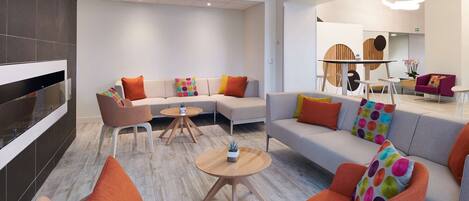 A modern hotel lobby with a sectional sofa, orange armchairs, and round tables.