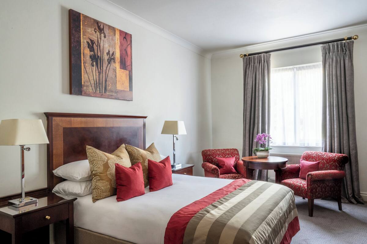 A hotel room with a large bed, two bedside tables with lamps, a small round table with a chair, and a window with curtains.
