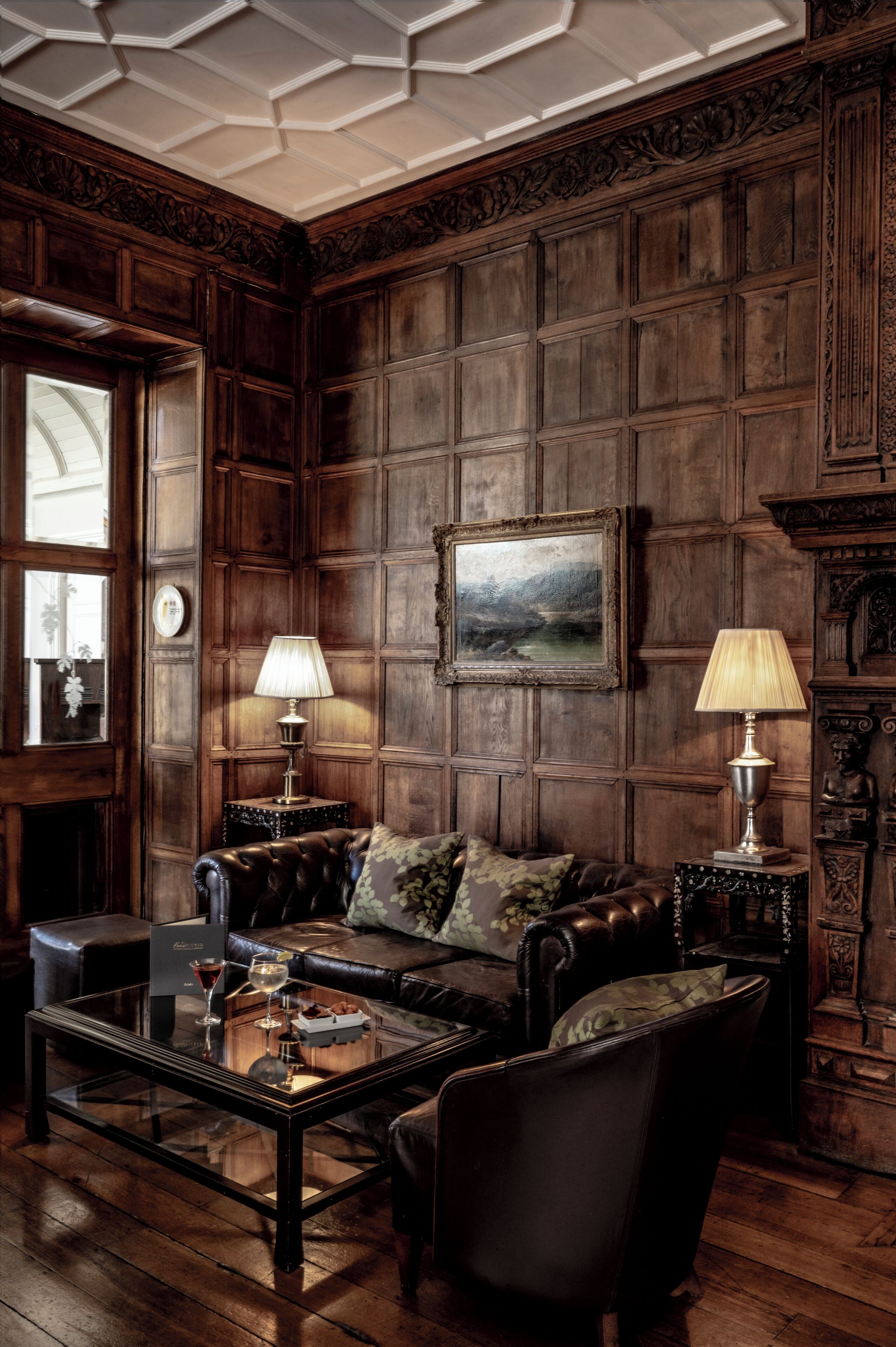 Una habitación con una amplia pared revestida de madera, un cuadro de paisaje enmarco, un sofá de cuero, dos lámparas y una mesa de centro de vidrio con una bebida y aperitivos.