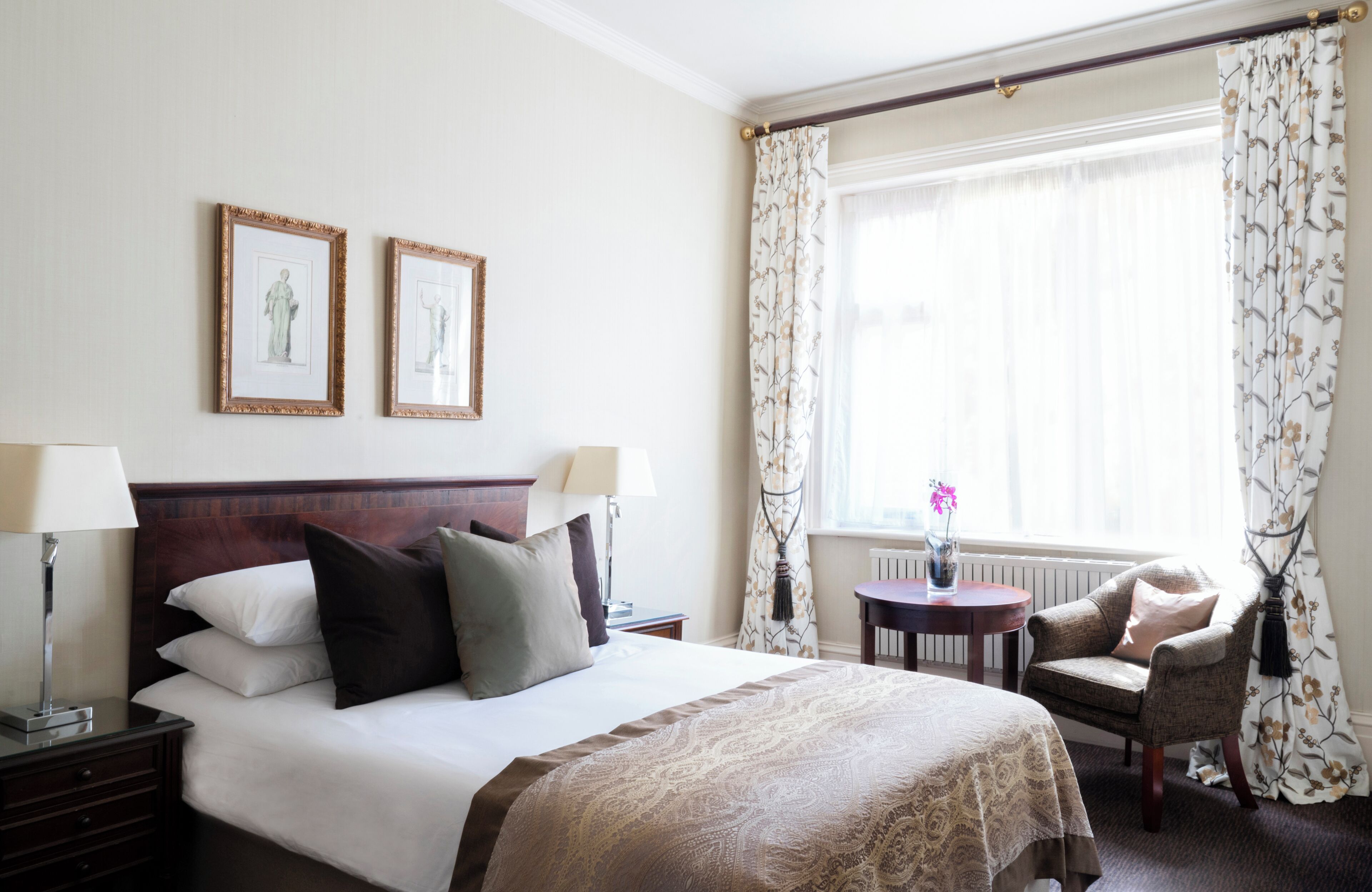 A hotel room with a large bed, two bedside lamps, a small table with a vase, a chair, and a window with curtains.