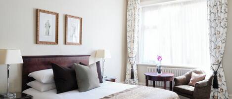 Une chambre d’hôtel avec un grand lit, deux lampes de chevet, une petite table avec un vase, une chaise et une fenêtre avec des rideaux.