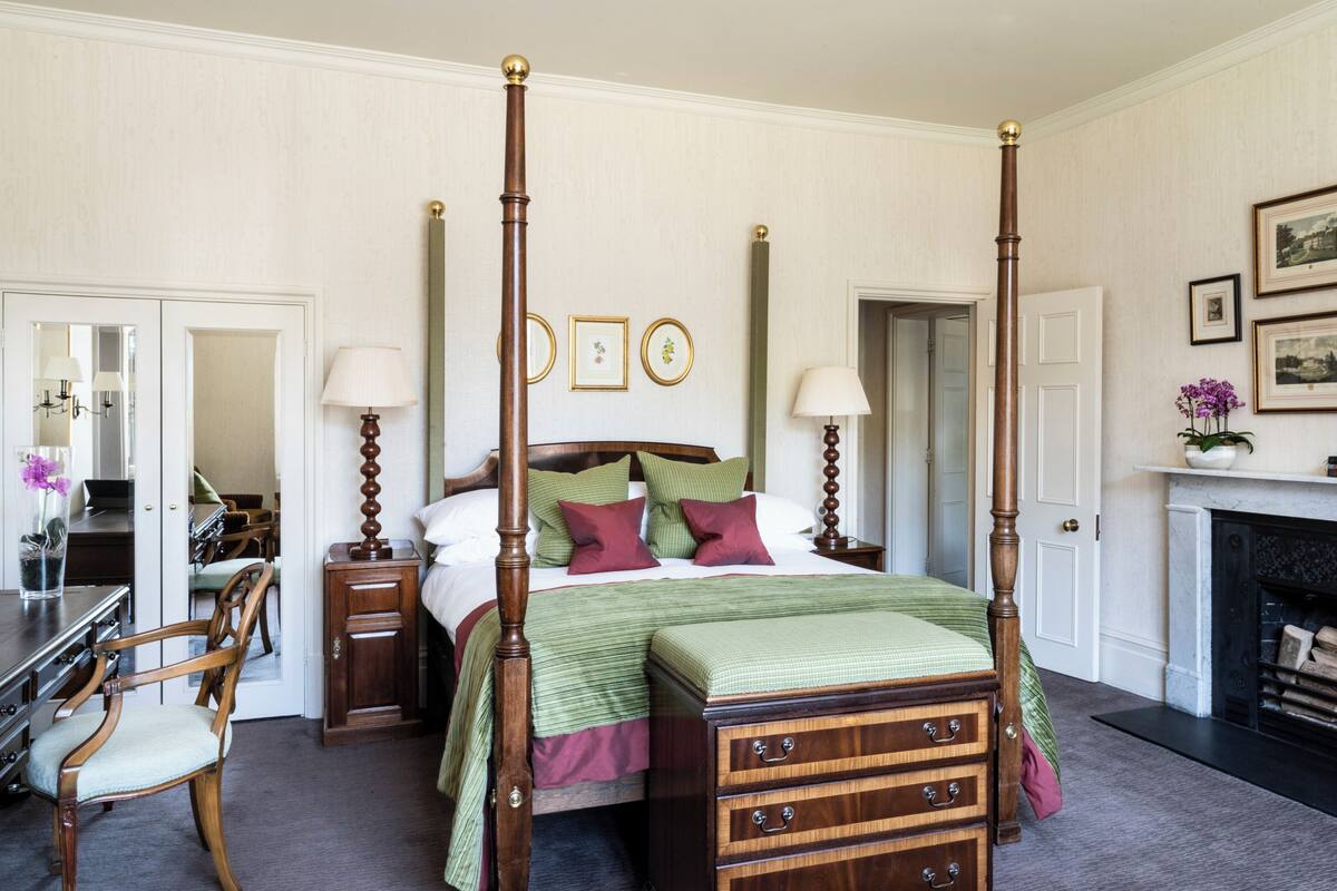 A four-poster bed with a green and red bedspread, wooden nightstands, a fireplace, and framed pictures on the wall.