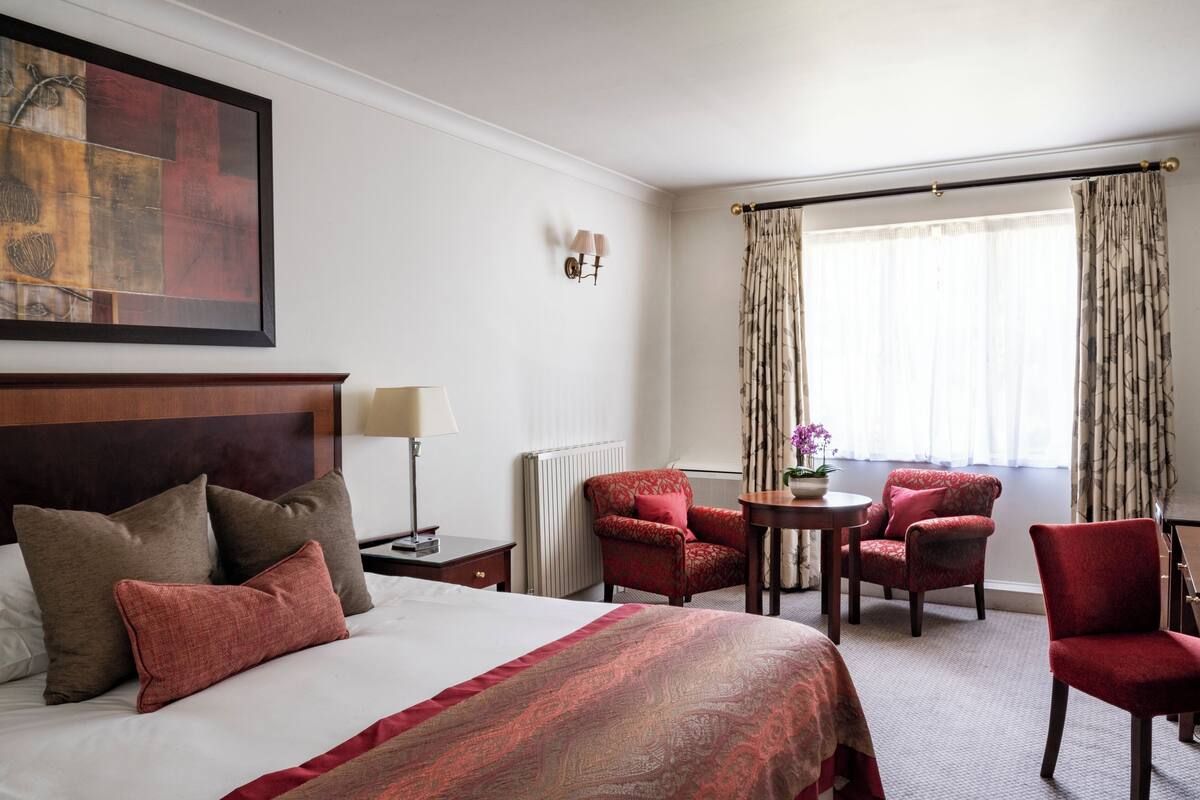 A hotel room with a large bed, two red armchairs, a small table with a vase, and a window with curtains.