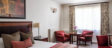 A hotel room with a large bed, two armchairs, a small table, a lamp, a painting on the wall, and a window with curtains.