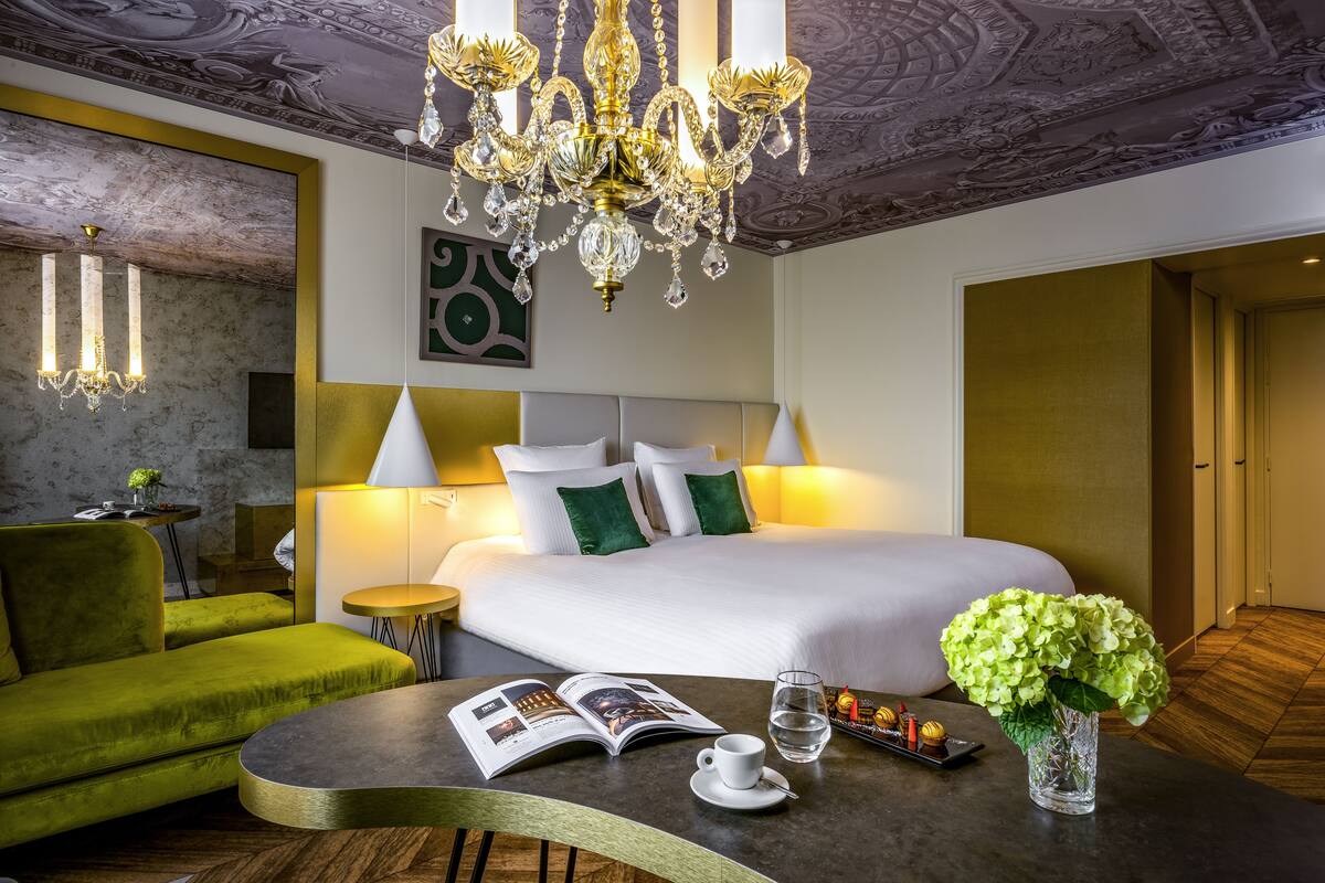 Une chambre d’hôtel avec un grand lit, un canapé vert et une table sur laquelle se trouvent un magazine, une tasse et des fleurs.