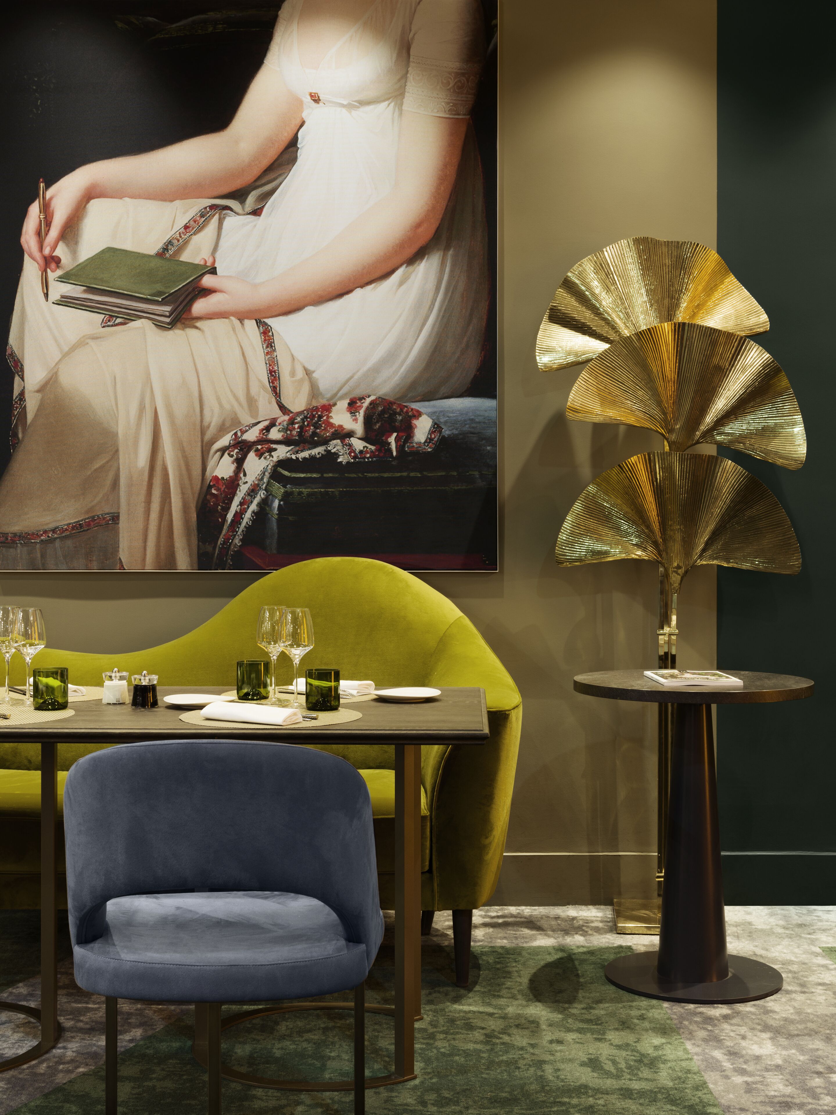 Un comedor con una mesa preparada para dos personas, una silla azul, un sofá verde y una lámpara decorativa.