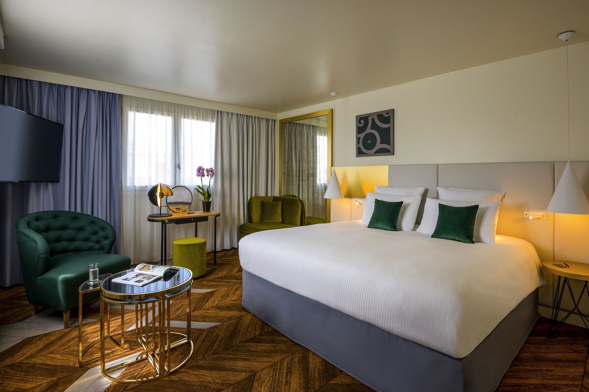 Een hotelkamer met een groot bed, twee fauteuils, een klein tafeltje, een televisie en een raam met gordijnen.