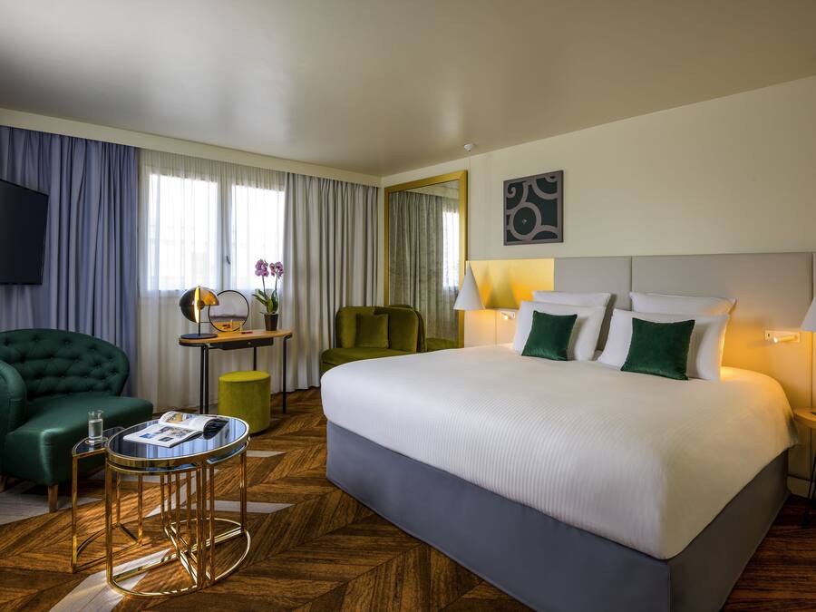 A hotel room with a large bed, two armchairs, a small table, a TV, and a window with curtains.