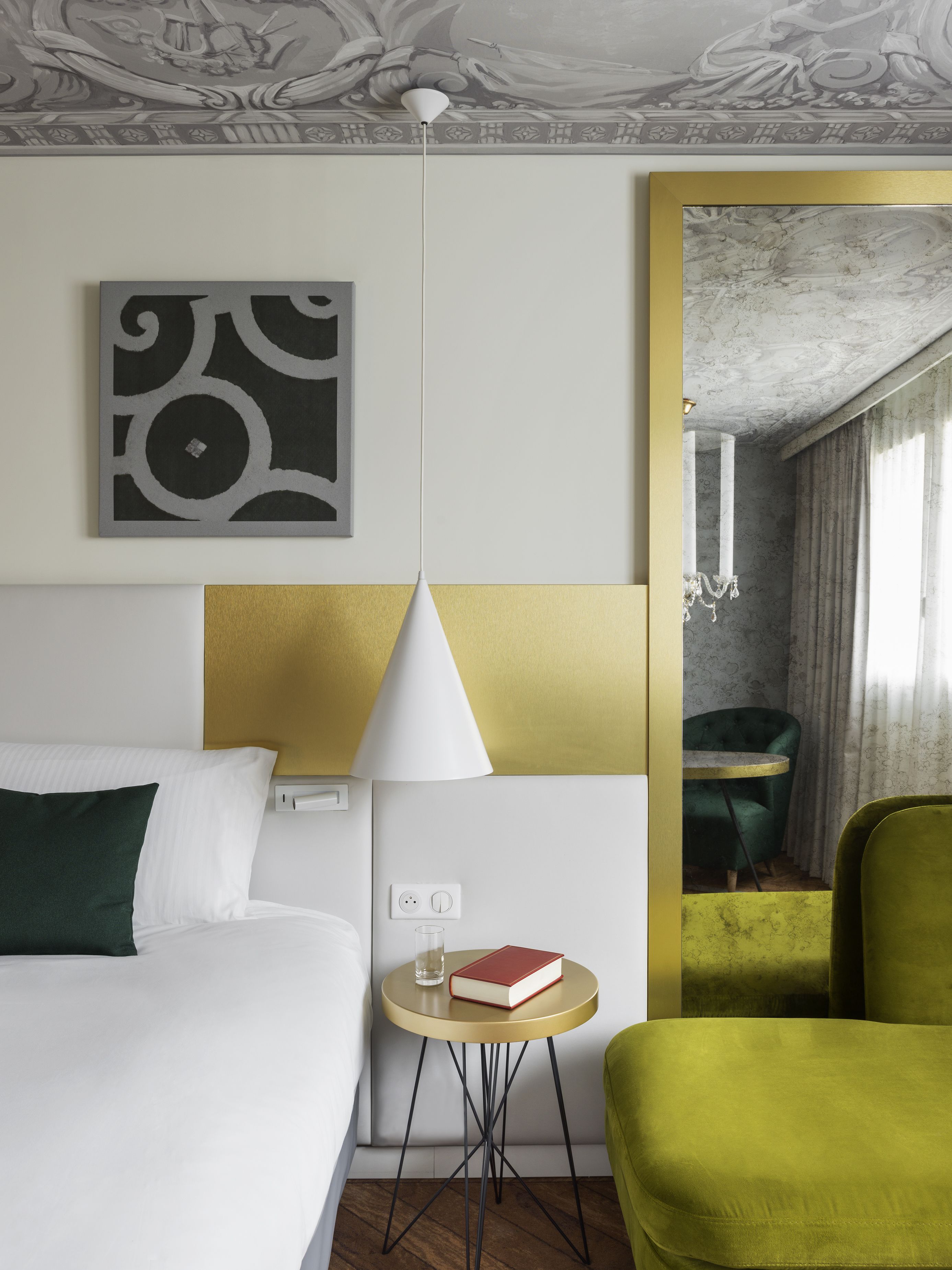 Une chambre d’hôtel avec un lit, une petite table ronde sur laquelle se trouvent un livre et un verre, un fauteuil vert et un miroir qui reflète un fauteuil vert et des rideaux.